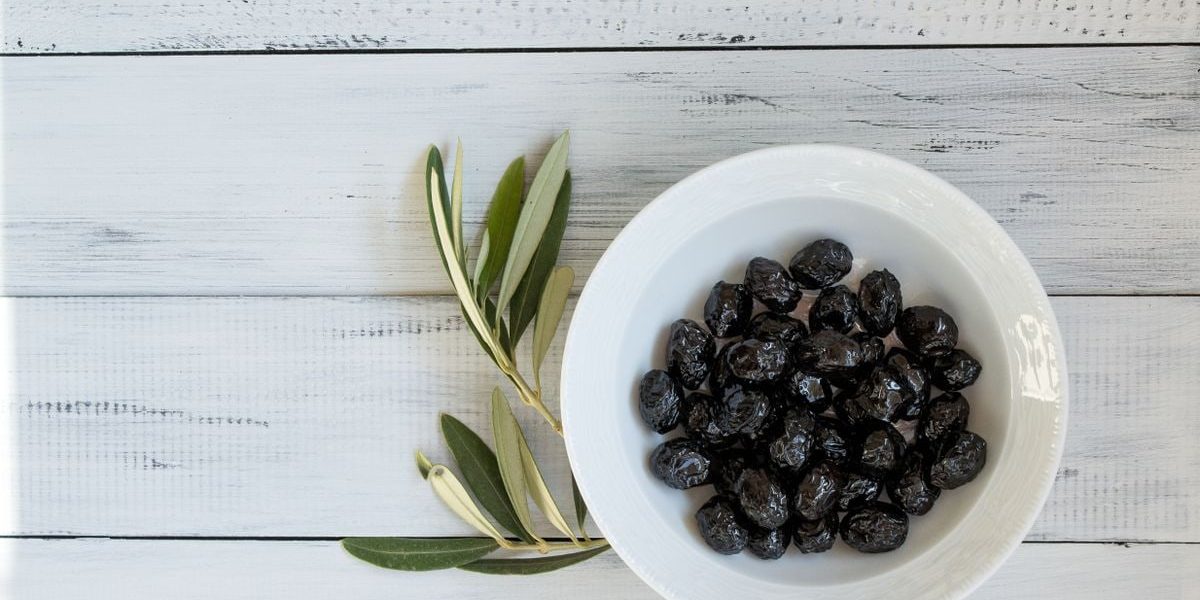 A bowl of black olives on a rustic white wooden surface with olive leaves. Perfect for culinary themes.