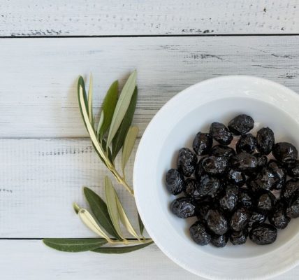 A bowl of black olives on a rustic white wooden surface with olive leaves. Perfect for culinary themes.