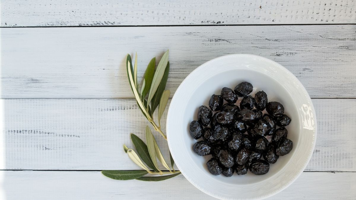 A bowl of black olives on a rustic white wooden surface with olive leaves. Perfect for culinary themes.